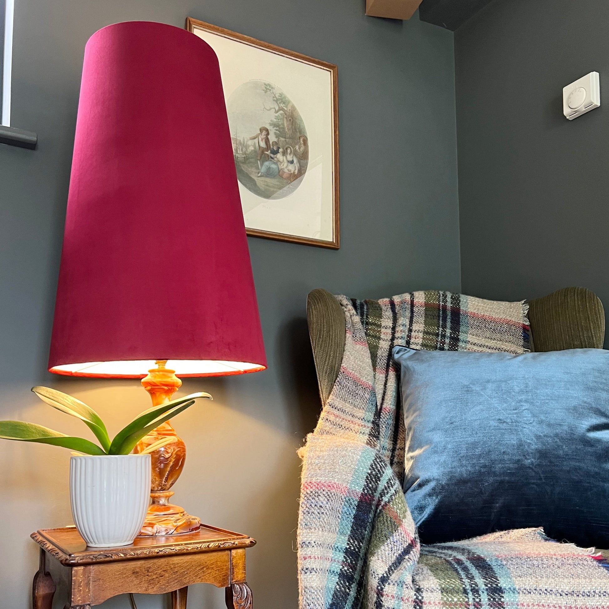 Chair with plaid blanket and blue pillow next to a lamp with a red shade on a wooden side table.